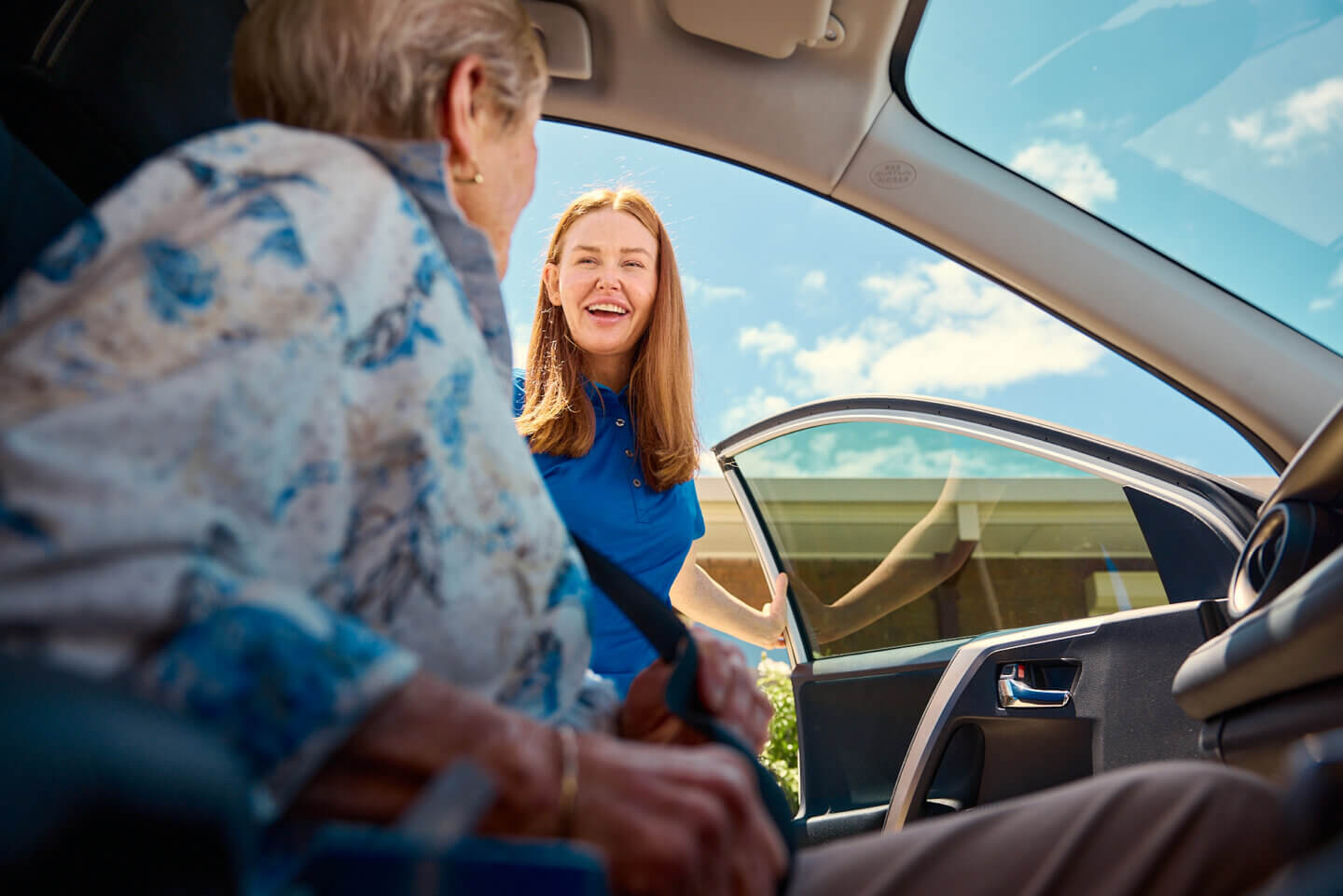 A smiling ECH worker in a blue shirt greets an elderly person as they sit in a car, viewed from inside the vehicle.